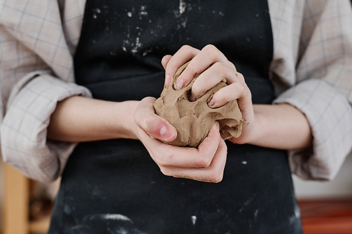 Hands of young creative woman in workwear holding piece of soft clay and kneading it while preparing for making earthenware products