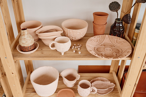 Assortment of handmade clay items on display consisting of wooden shelves by wall in small souvenir shop or pottery workshop