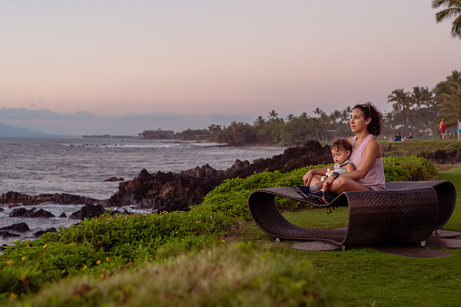 A young multiracial woman affectionately holds her toddler daughter in her lap while sitting on a bench at the beach and watching the sunset.