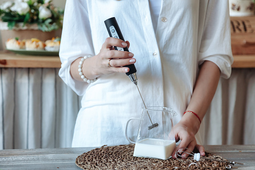 Mixing milk in glass pot by frother close up. Woman making foamy milk with handheld mixer. Housewife female in home clothes using mixer whips cooking food in kitchen at home. Cooking lifestyle concept