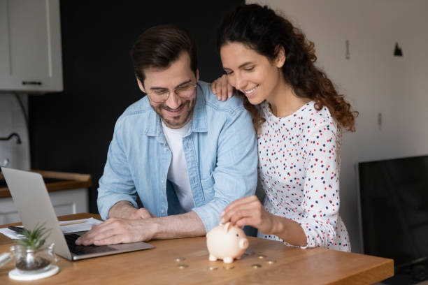 Couple in domestic kitchen with laptop putting coin in piggybank stock photo