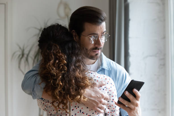 Jealous husband hugging wife and checking her phone stock photo