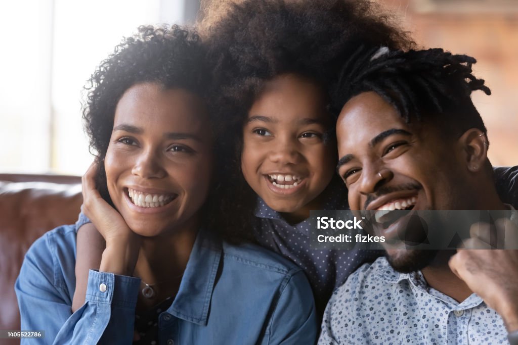 Happy bonding African American family watching smart TV. Head shot happy bonding African American two generations family parents resting with little cute child daughter on cozy sofa, looking in distance, watching funny smart TV programs or shows at home. Afro Hairstyle Stock Photo