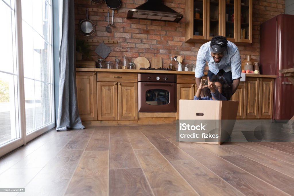 Cheerful African family having fun in kitchen. Cheerful young African ethnicity father pushing seated in carton box laughing little biracial kid daughter, having fun involved in entertaining pirates game leisure activity together in kitchen. Foster Care Stock Photo