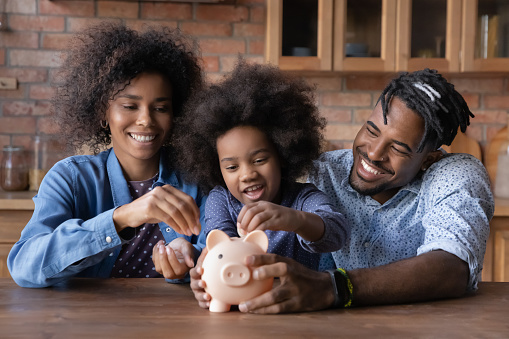 Caring young African American couple parents teaching small biracial kid daughter saving money, planning future purchases together, putting coins in small piggybank, financial education for children.