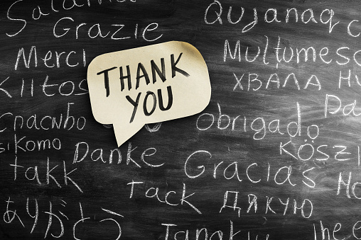 Thank You in many languages written with chalk on blackboard. The English translation is written on a adhesive note cut in form a speech bubble.