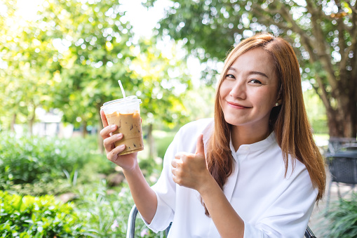 Portrait image a young asian woman holding and making thumbs up hand sign to a cup of iced coffee in the outdoors