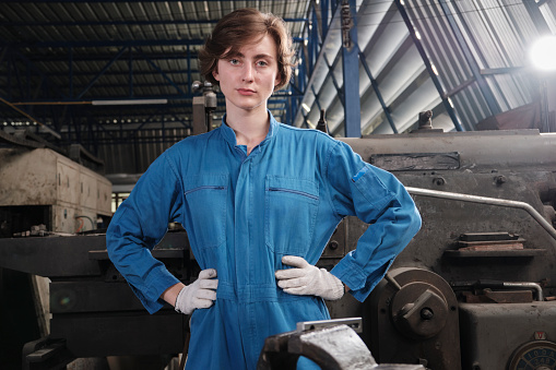 Portrait of one young Caucasian female industry worker in safety uniform stand, looking at camera and akimbo and expresses solemn confidence in a mechanical factory. Professional engineer occupation.