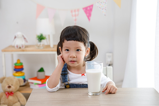 Little asian girl sitting at table in room, Preschooler girl drinking some milk with glass on sunny day, kindergarten or day 
care. Educational concept for school kids.