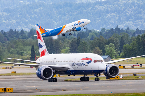 Portland, Oregon, USA - June 12, 2022: A British Airways Boeing 787 Dreamliner taxies to the gate after a nine hour direct flight from London, England. Taking off in the background is an Allegiant Air A320.