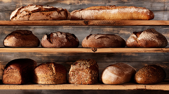 Bunch of assorted freshly baked bread loaves with different shapes and baguettes placed on shelves against wooden background in light studio