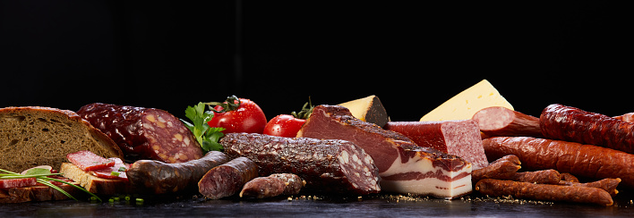 Assorted types of cured sausages neat bread placed on dark surface with vegetable and cheese in studio on black background