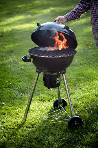 Crop unrecognizable male near kettle charcoal barbecue grill with black lid and burning hot fire placed on grassy lawn in countryside