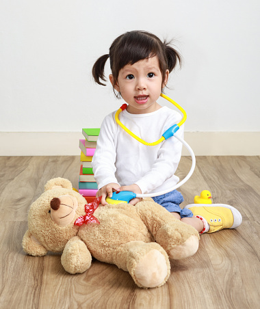 Asian little doctor girl play with toy teddy bear in the room.