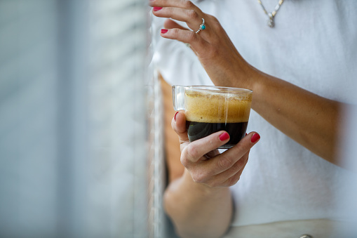 Close up shot of unrecognizable woman standing by the window, peeking through the blinds and checking the weather outside while enjoying a cup of morning coffee.