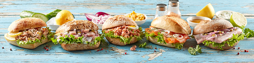 Row of traditional German Fischbrotchen sandwiches with various types of fish and shrimps with green lettuce served with sauces on blue wooden table