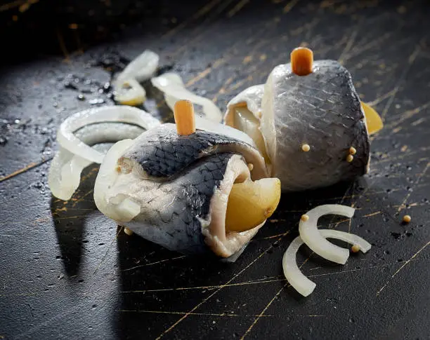 Closeup of traditional German dish rollmops made with marinated herring fillet with sliced onion and lemon on black surface