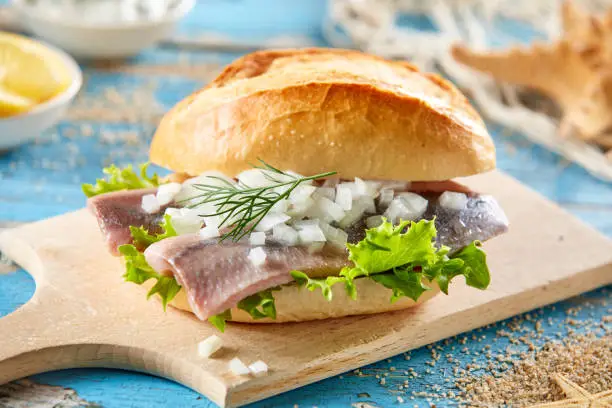 Yummy sandwich with pickled herring and chopped fresh onion served on wooden cutting board on blue table decorated in marine style