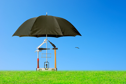 Tool House protected by black umbrella in meadow against blue sky,\nInsurance and real estate concept.