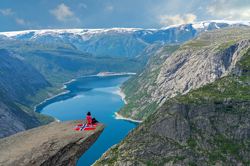 Norway - June 27, 2022: Rock landscape, Norway. Trolltunga rock. Girl sitting with Norwegian flag. Mountain lake Ringedalsvatnet landscape.