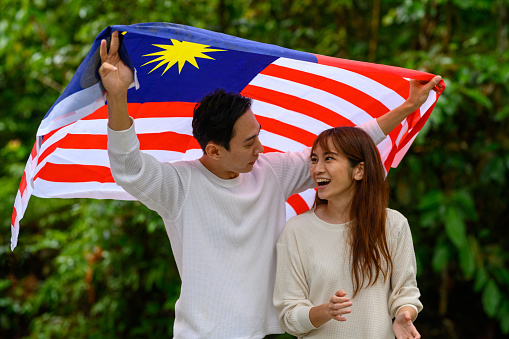 Young Asian Couple feeling cheerful spending leisure time at public park holding Malaysian flag to celebrate Independence Day.