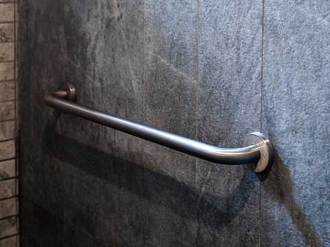 Close-up photo of stainless steel grab bar handrail installed on grey stone tiles wall background in a hotel handicapped disabled access bathroom.
