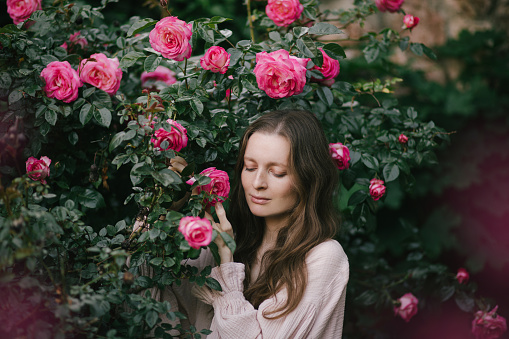 Young beautiful woman wearing pink dress enjoying fragrance of blooming roses in a spring garden.
