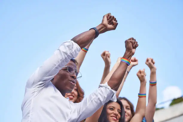 Group of multiracial people raising fists with LGBT rainbow flag wristbands outdoors. Diversity, equality and unity concept.