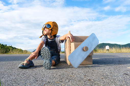 Cute dreamer little girl playing with cardboard planes on a lake road on a sunny day. Happy kid playing with cardboard plane against blue summer sky background. Childhood dream imagination concept.