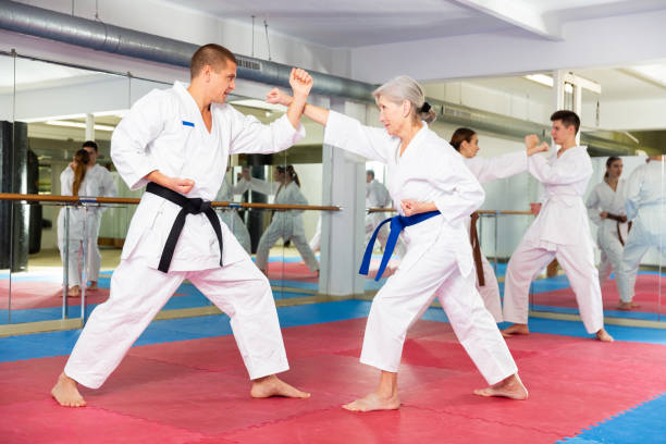 Aged woman in white kimono sparring with male opponent in gym Portrait of concentrated aged woman wearing white kimono sparring with male opponent during martial arts training in gym taekwondo stock pictures, royalty-free photos & images