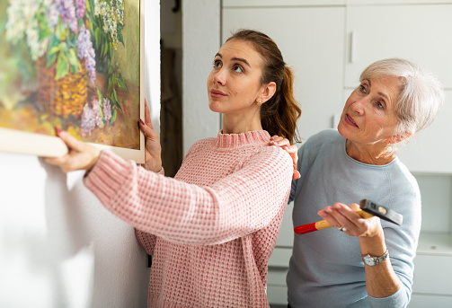 Young girl and older woman hanging together painting with depicted bouquet of lilacs on wall indoors