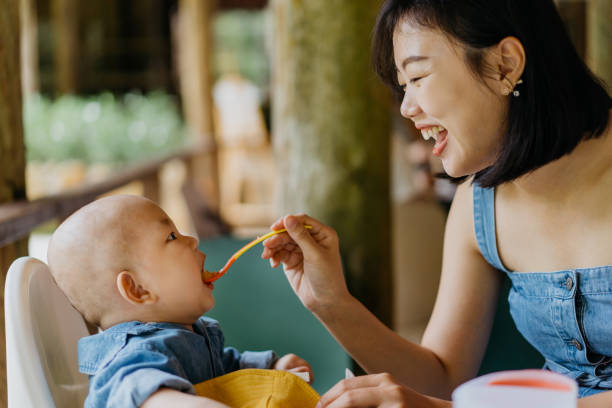Asian woman spoon feeding rice cereal to her baby boy Image of an Asian Chinese mother feeding rice cereal to her baby boy with baby spoon in a cafe baby spoon stock pictures, royalty-free photos & images