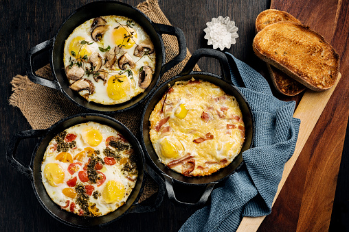 Flat-lay view of 3 baked egg recipes ready just out of the oven. From the top, eggs with mushrooms and thyme, next we have  eggs with ham and cheese, the third dish is eggs with tomatoes and fresh pesto.