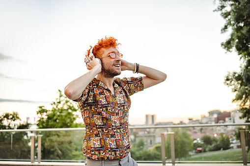 An alternative-looking redhead man standing on the balcony and listening to music