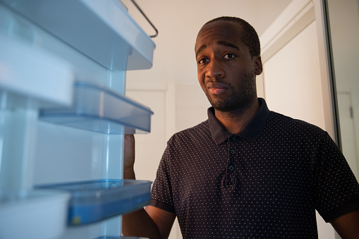 An African American man opens the fridge and realizes it is empty. He has a sad and worried facial expression. Crisis and poverty concept.