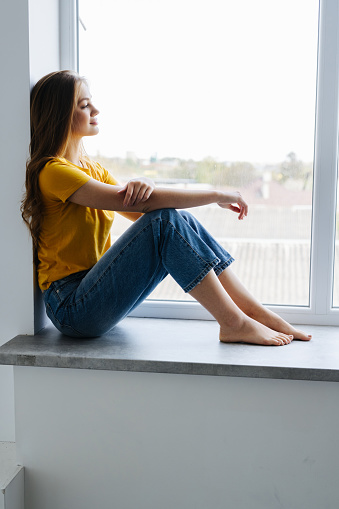 Young woman looking through the window with a city view, sitting on a windowsill in the morning.
