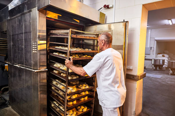 istock Professional baker in uniform inserts cart with decks for baking raw dough to make bread in an industrial oven in a bakery 1405581737