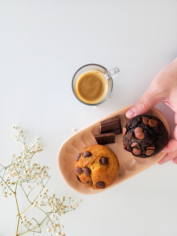 hand taking a cup of coffee and vanilla and chocolate muffins on pink plate on white background