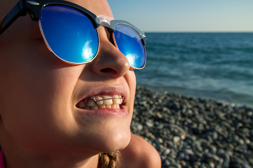 A girl with a removable orthodontic appliance and in sunglasses sits on the seashore. Concept of pediatric dentistry, correcting the bite.