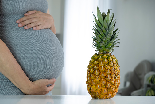 Pregnant woman with big belly and big fresh pineapple at home interior.