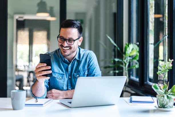 istock Cheerful freelancer with laptop text messaging on smart phone at home office 1405587994