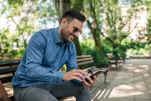 Smiling young businessman networking on smart phone while sitting on bench at park