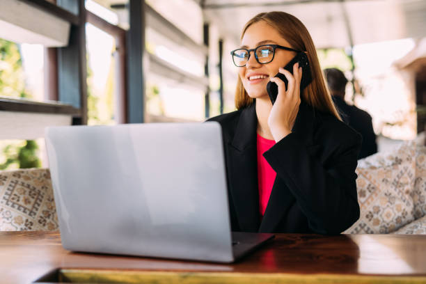 dreaming attractive girl in trendy blouse talking by mobile phone, businesswoman looking up, while sitting with laptop at the cafe, portrait of woman outdoors. - looking up young women outdoors laptop imagens e fotografias de stock