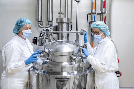 Two female employees in a laboratory wearing protective gloves, mask, cap and suit seen standing by the machine that is the part of the medicaments production during the working hours in a pharmaceutical manufacturing.