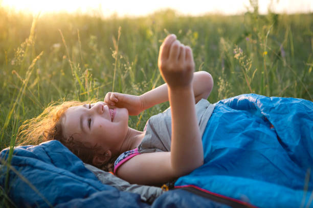 the girl is dissatisfied with scratching mosquito bites, child sleeps in a sleeping bag on the grass in a camping trip. eco-friendly outdoor recreation, summer time. sleep disturbance, repellent. - mosquito child bug bite scratching imagens e fotografias de stock