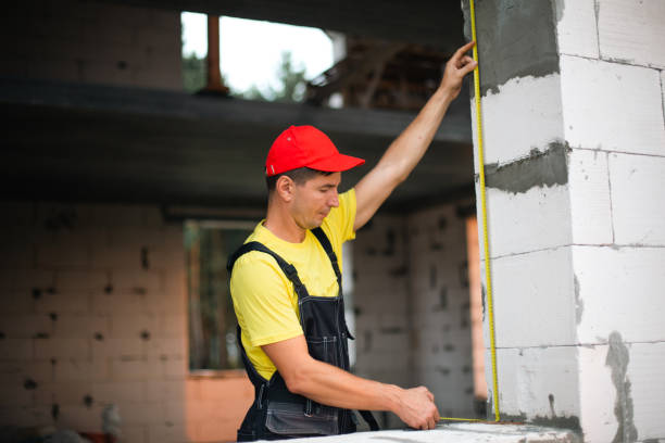 istock Construction worker at construction site measures the length of window opening and brick wall with tape measure. Cottage are made of porous concrete blocks, work clothes - jumpsuit and baseball cap 1405595664