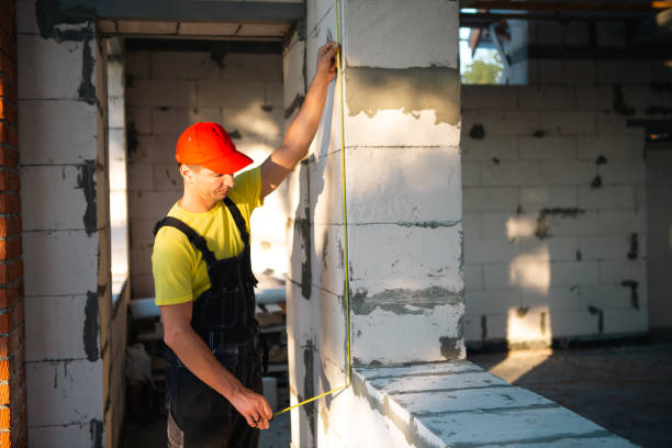 istock Construction worker at construction site measures the length of window opening and brick wall with tape measure. Cottage are made of porous concrete blocks, work clothes - jumpsuit and baseball cap 1405595687