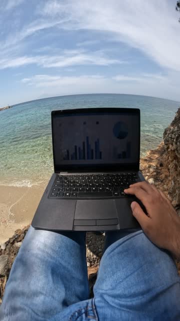 Young man working on laptop on the vacation at the beach. Finishing up work to relax. His POV.