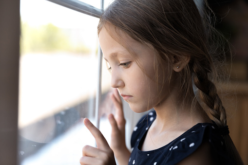 Sad thoughtful orphan child got trauma, feeling depressed, lonely, upset. Serious bored girl standing by window, thinking, touching glass. Childhood problems, abuse, violence in family concept