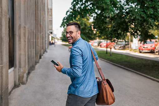 Cheerful businessman with smart phone and bag looking over shoulder while walking on sidewalk in the city